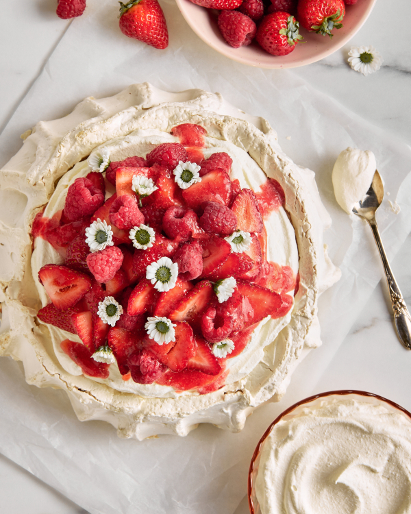 Vue de dessus d'une Pavlova garnie de framboises, de fraises et de fleurs, présentée avec un bol de baies, un bol de crème fouettée et une cuillère dorée.