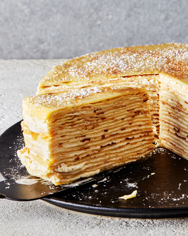 Un gâteau crêpe au bourbon saupoudré de sucre glace sur un plateau noir, une tranche étant retirée à l'aide d'un lève-tarte.