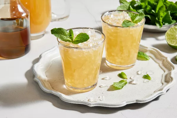 Deux verres de julep à la menthe sans alcool avec de la glace pilée et garnis de menthe, servis sur un plateau blanc et présentés avec un bol de feuilles de menthe, une moitié de lime, une carafe de julep préparé et une carafe de sirop simple à la menthe.