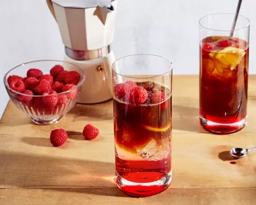 Deux verres d'espresso tonique glacé à la framboise sur une table en bois, accompagnés d'un bol de framboises et d'une machine à