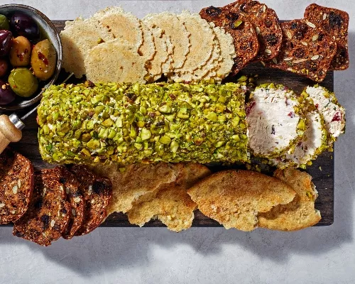 Une bûche au fromage à la crème enrobée de pistaches concassées sur une planche avec croustilles, craquelins et olives